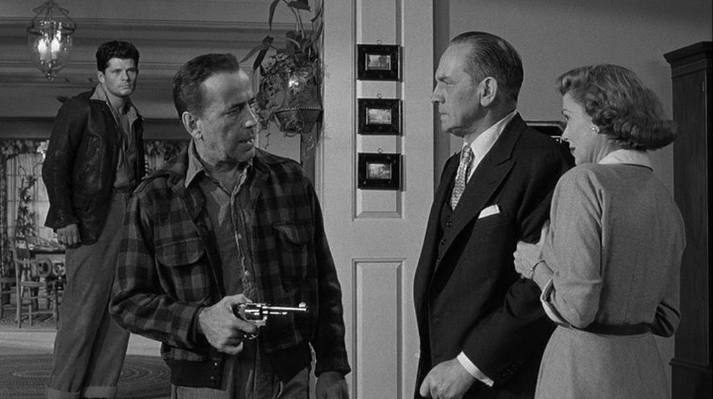 Black-and-white scene: Humphrey Bogart as desolate escapee Glenn Griffin, who threatens the Hilliard couple, played by Fredric March and Martha Scott, in their living room with a revolver; in the background, Griffin's accomplice and younger brother Hal Griffin, played by Dewey Martin, looks on.