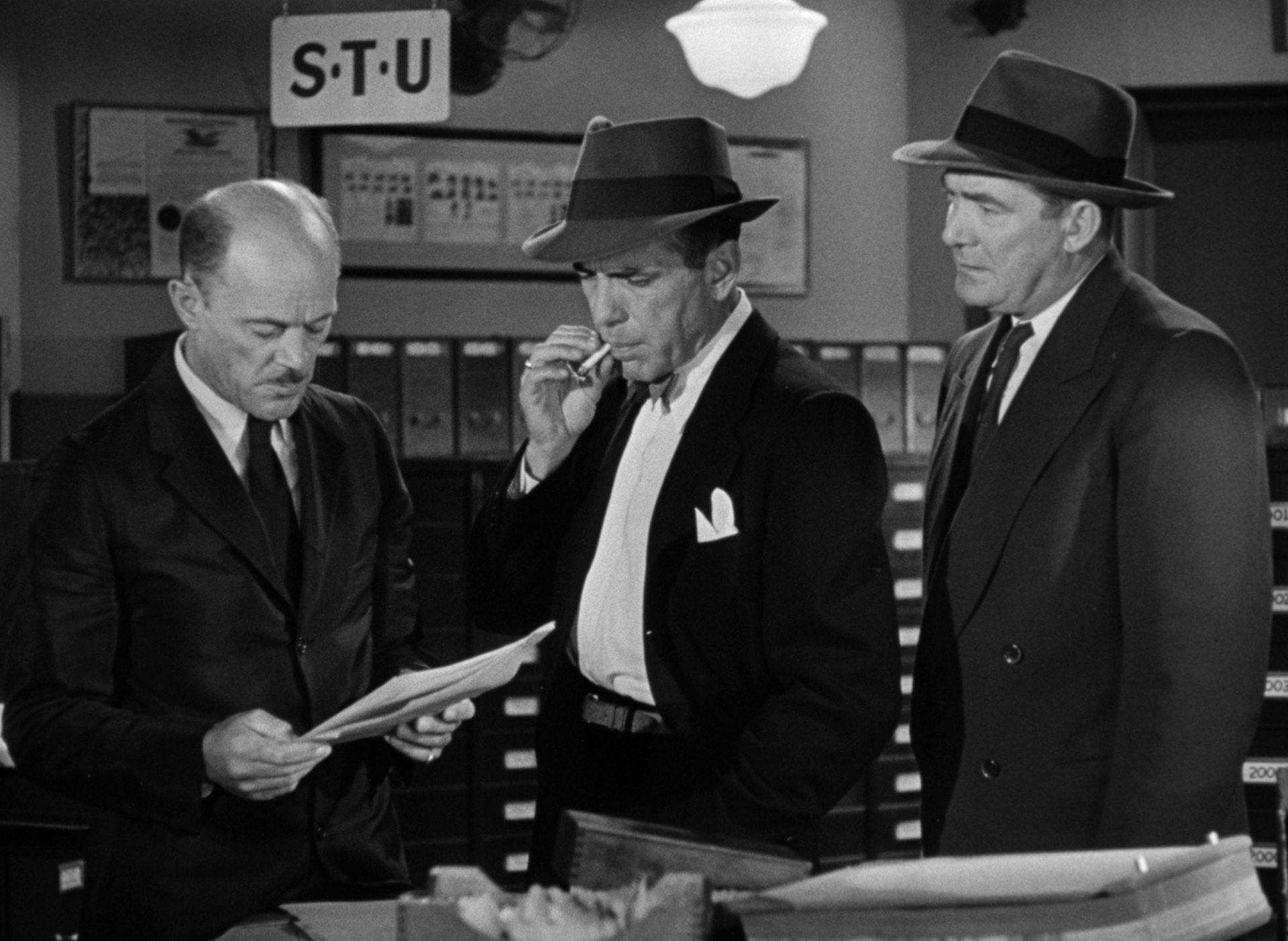 Black-and-white scene in an office: Humphrey Bogart as a prosecutor accompanied by a cop talking to an employee who is reading a file; Bogart is wearing a fedora and taking a drag on his cigarette.