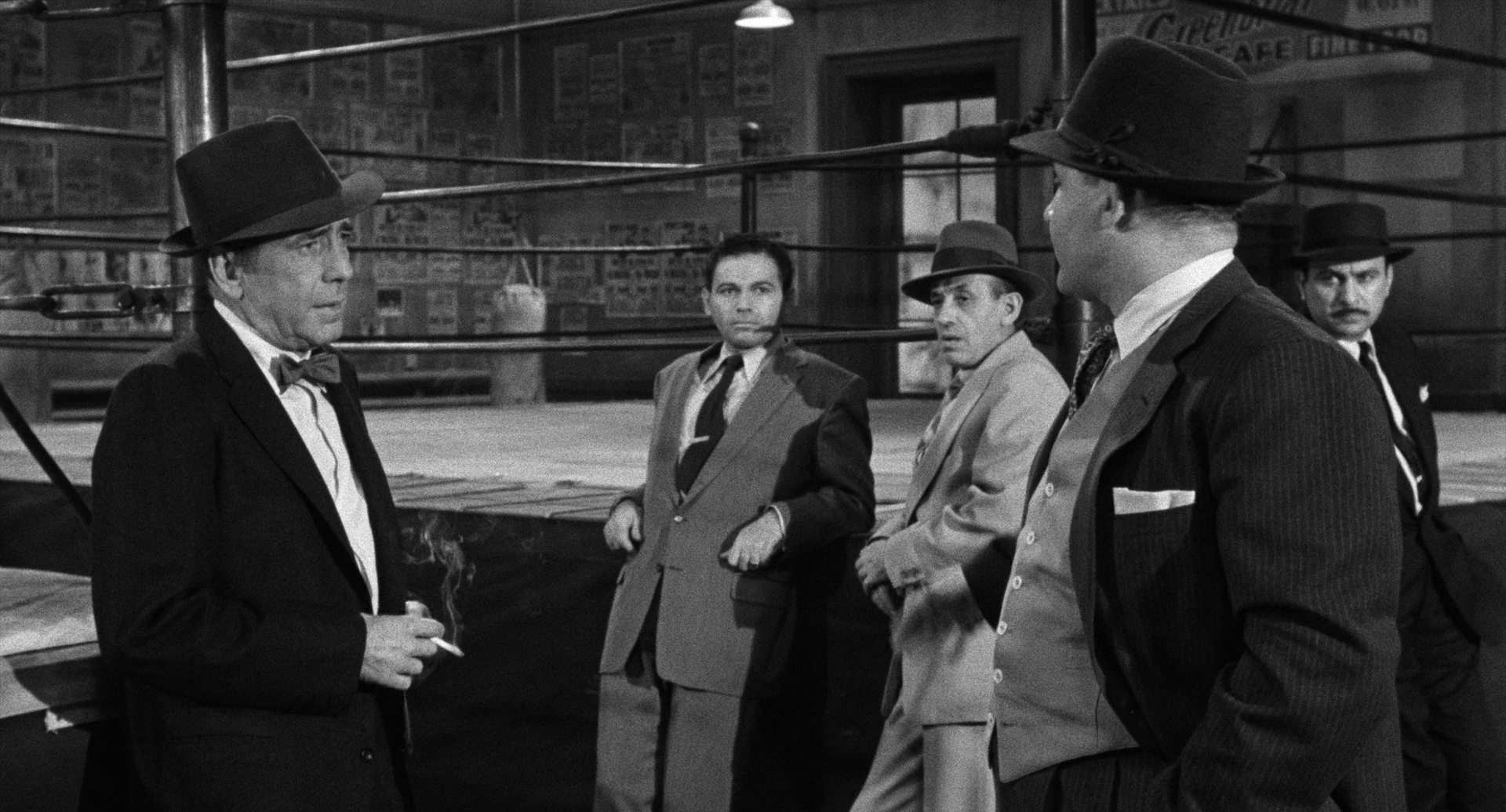 Black-and-white scene: Humphrey Bogart, wearing a hat, bow tie and holding a cigarette, stands at the boxing ring on the left of the picture, talking to Rod Steiger as a boxing promoter; there are also three dodgy men at the ring.