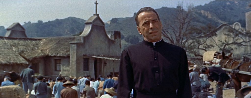 Humphrey Bogart with a serious look in the garb of a priest against the backdrop of a small church, in front of which there is a crowd of people, and an Asian mountain landscape.
