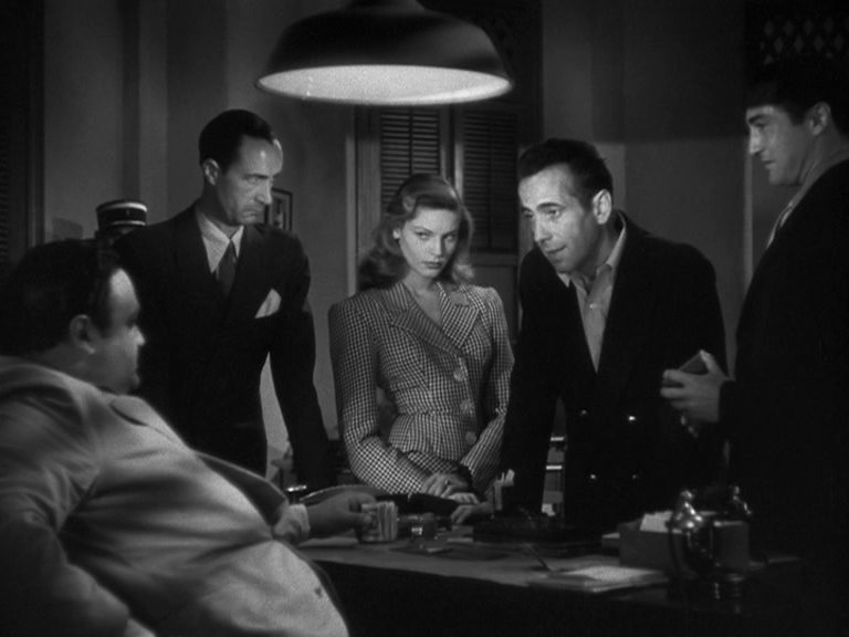 Schwarz-Weiß-Szene mit Lauren Bacall und Humphrey Bogart – beide stehen, umringt von zwei Männern, vor einem Schreibtisch, an dem ein Mann sitzt; Bogarts Figur starrt selbstbewusst, Bacalls Charakter blickt mysteriös.