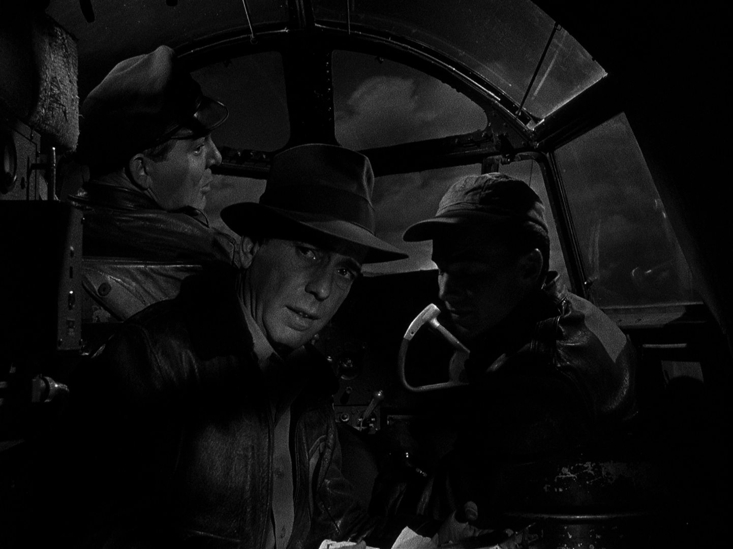 Black and white scene with a nighttime look into the cockpit of a cargo plane: Humphrey Bogart as Joe Barrett in Indiana Jones-like clothing in the presence of the two pilots.