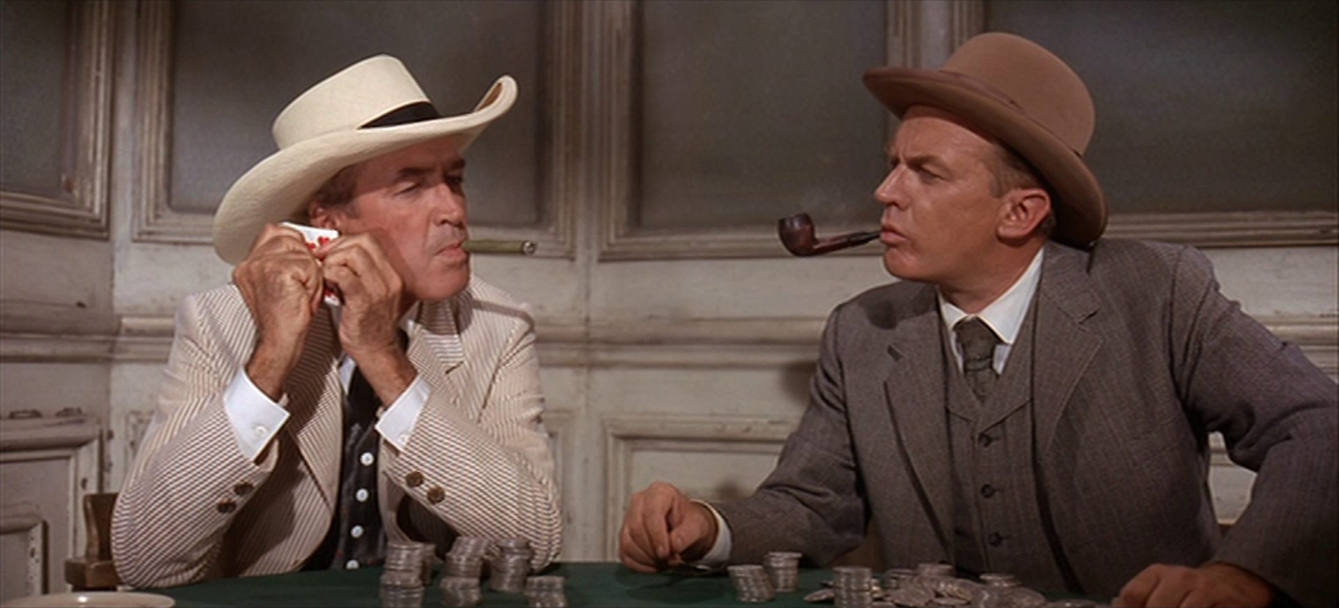 Nahaufnahme von Wyatt Earp und Doc Holliday am Pokertisch in einem Saloon; Earp raucht eine Zigarre und hält sich mit konzentriertem Gesicht einen Stapel Spielkarten an sein rechtes Ohr; Holliday raucht Pfeife und beobachtet Earp; vor ihnen sind ihre Pokereinsätze aufgetürmt.