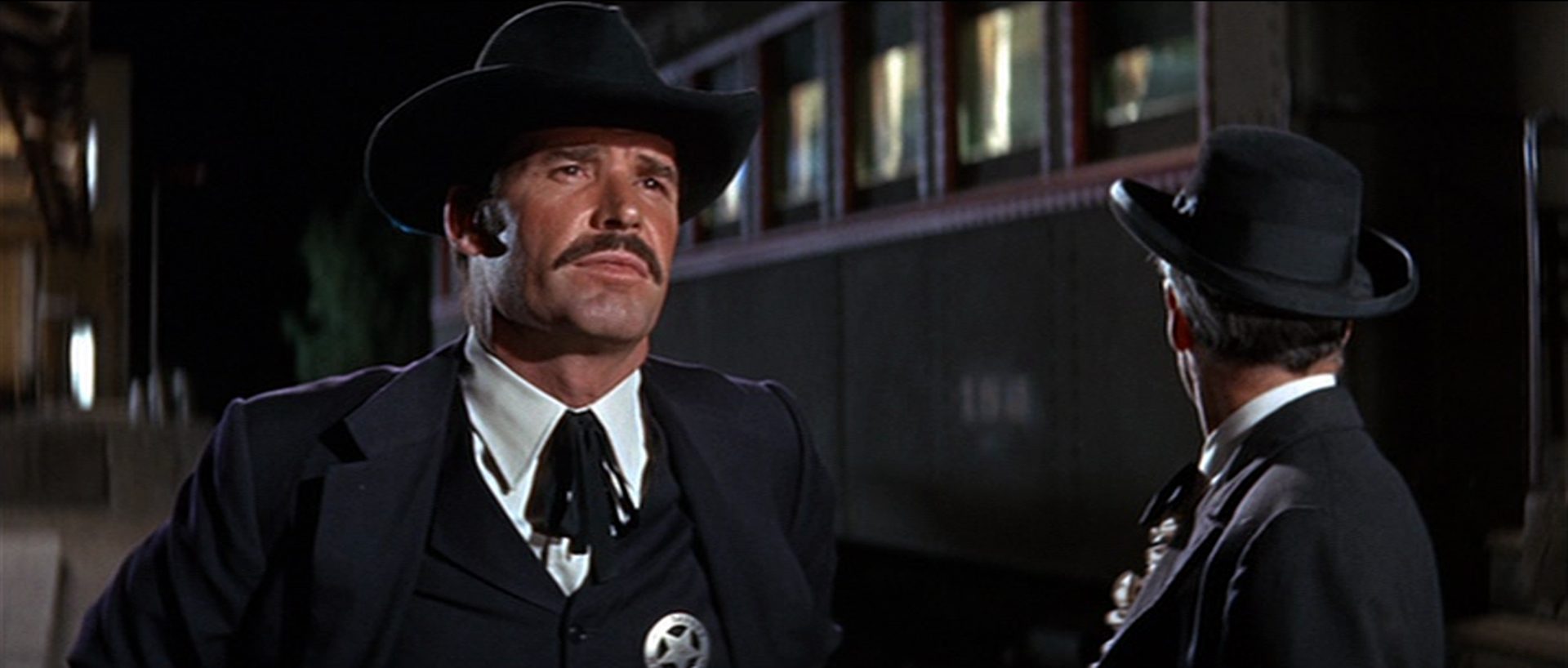 Nahaufnahme von Wyatt Earp mit Schnauzbart und festem Blick vor einem Eisenbahnwaggon, neben ihm Doc Holliday.