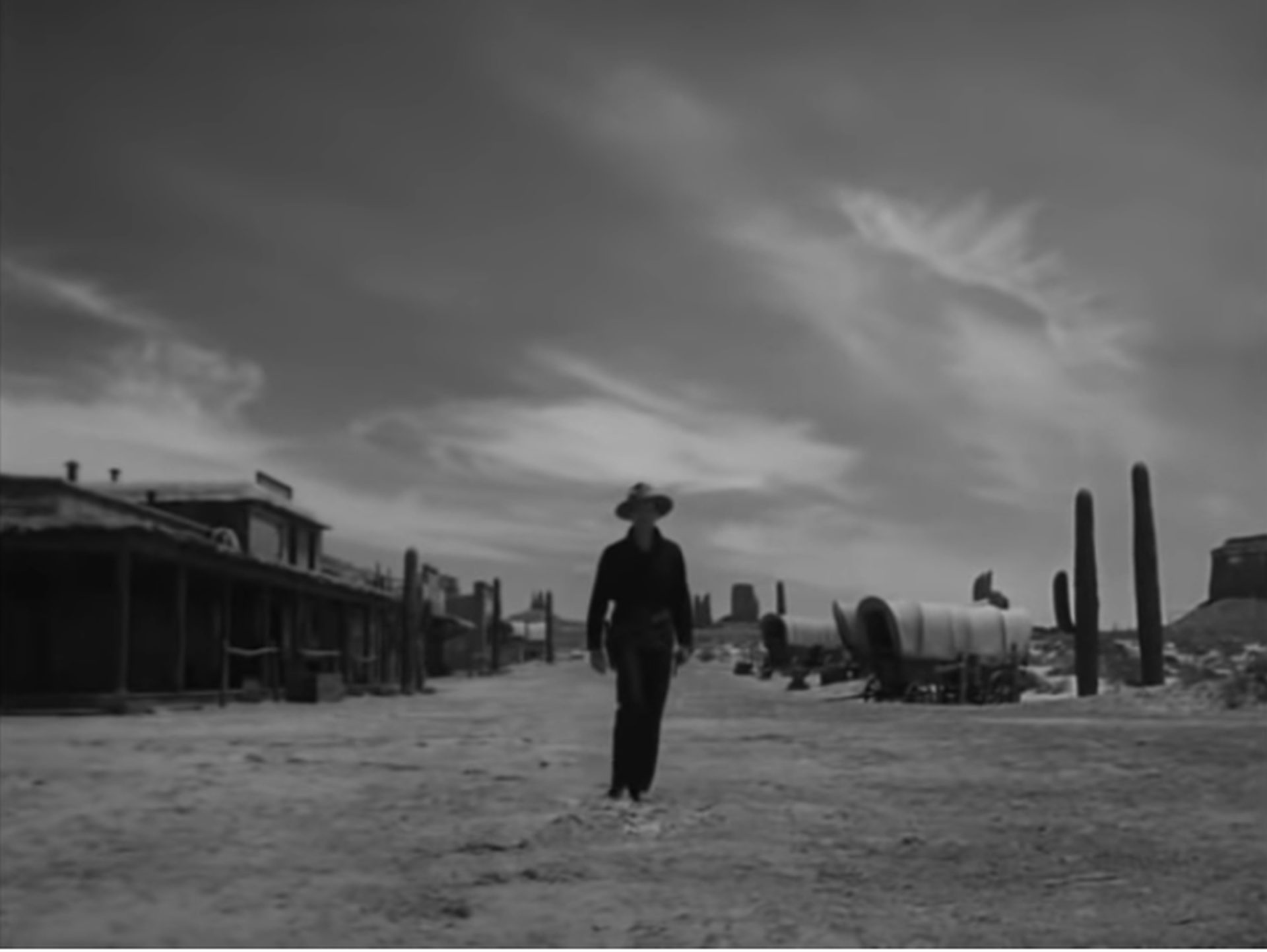 Wyatt Earp schreitet allein auf die Kamera zu, im Hintergrund mit spektakulärem Himmel befinden sich die flachen Gebäude einer Kleinstadt, Planwagen und Felsen des Monument Valley