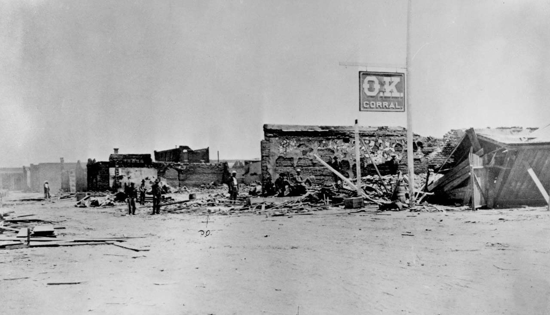 Schwarz-Weiß-Aufnahme des originalen „O.K. Corral“ als Ruine mit eingestürzter Scheune und verbrannter Mauer. Lediglich das Schild mit der Aufschrift „O.K. Corral“ ist intakt.