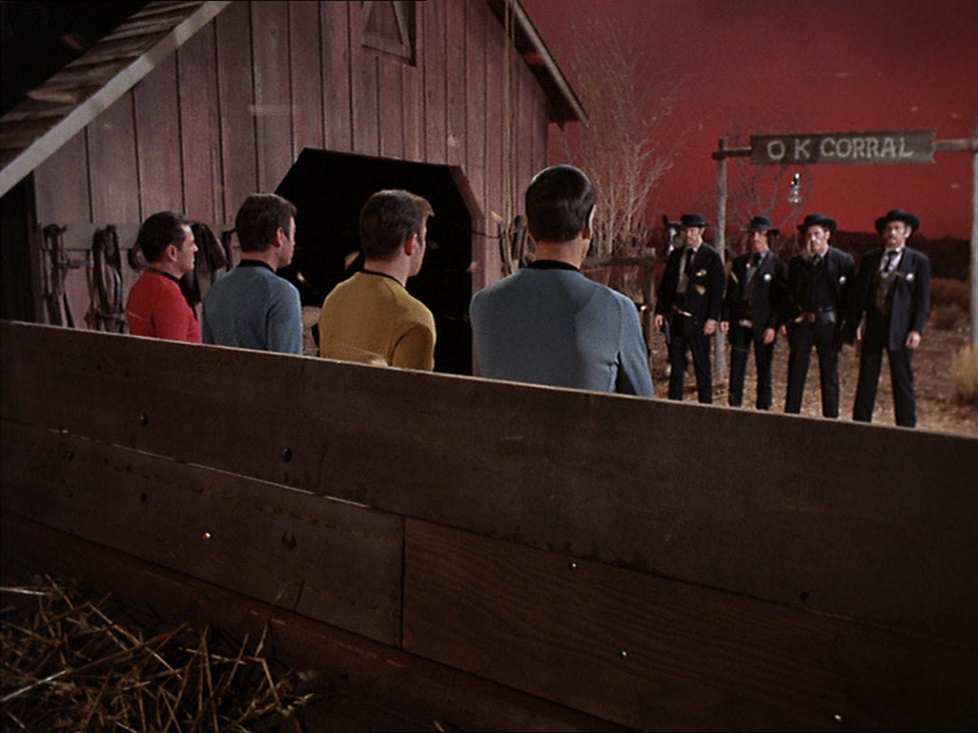 Vier Mitglieder der „Enterprise“-Crew mit dem Rücken zur Kamera; im Hintergrund die Earps mit Doc Holliday unter einem Schild, auf dem „O K Corral“ geschrieben steht.
