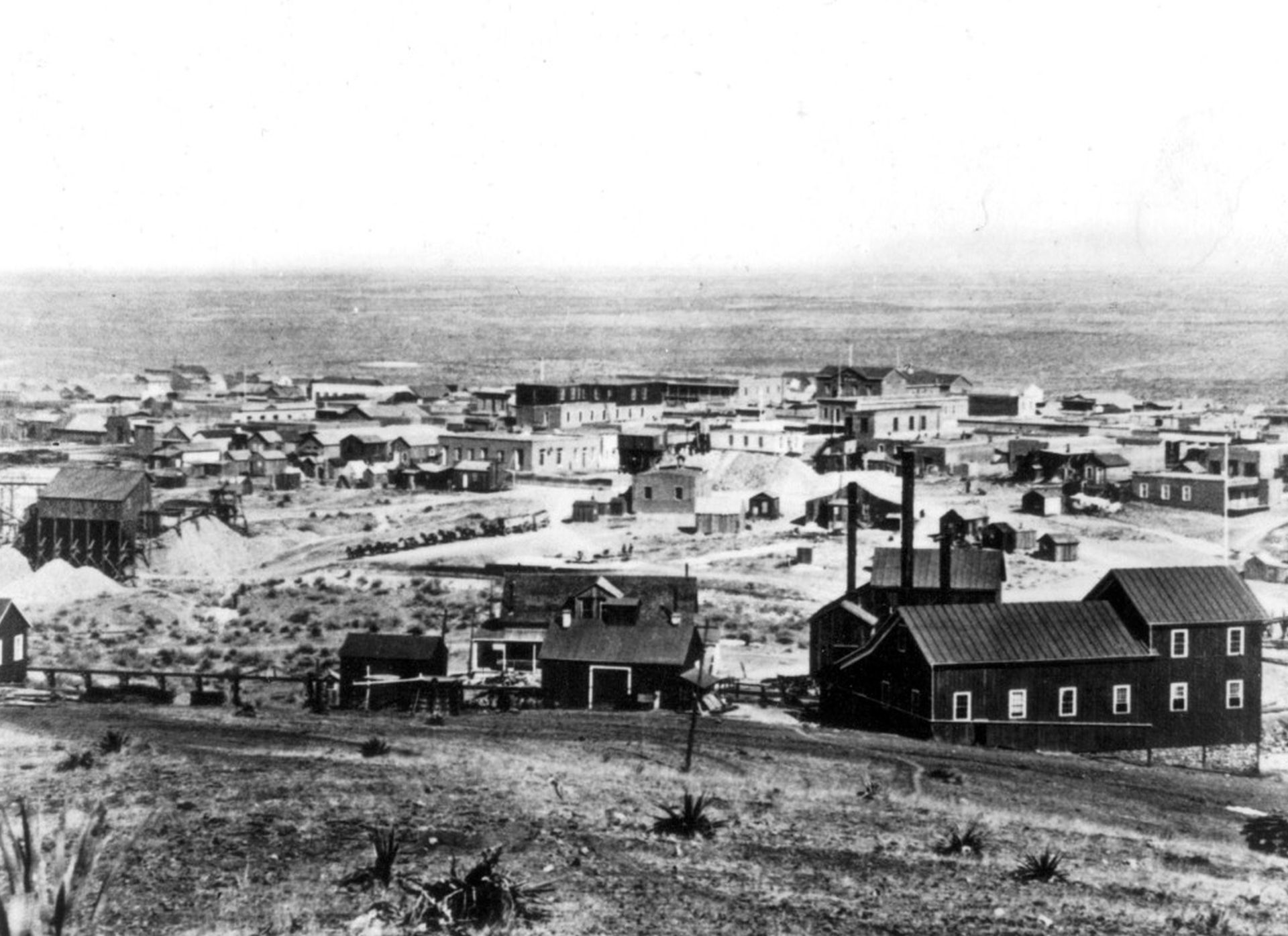 Schwarz-Weiß-Aufnahme mit Blick auf die vorwiegned aus einfachen Holzgebäuden bestehende Stadt von einer Anhöhe aus.