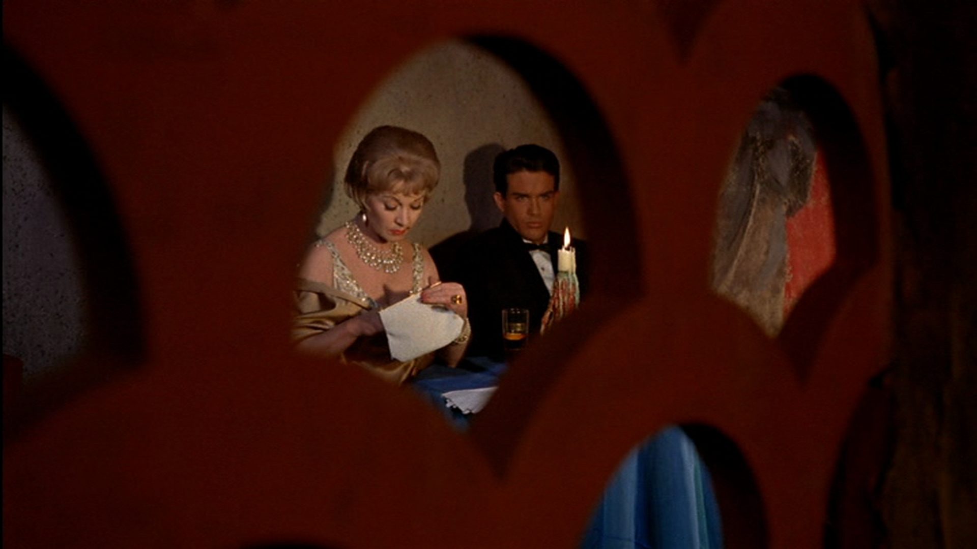 Vivien Leigh und Warren Beatty in einem italienischen Etablissement durch eine kleine Öffnung einer roten Abtrennung betrachtet; sitzen an einem kleinen Tisch bei Kerzenlicht; sie sucht etwas in ihrer Handtasche, er starrt angestrengt in den Raum hinein.