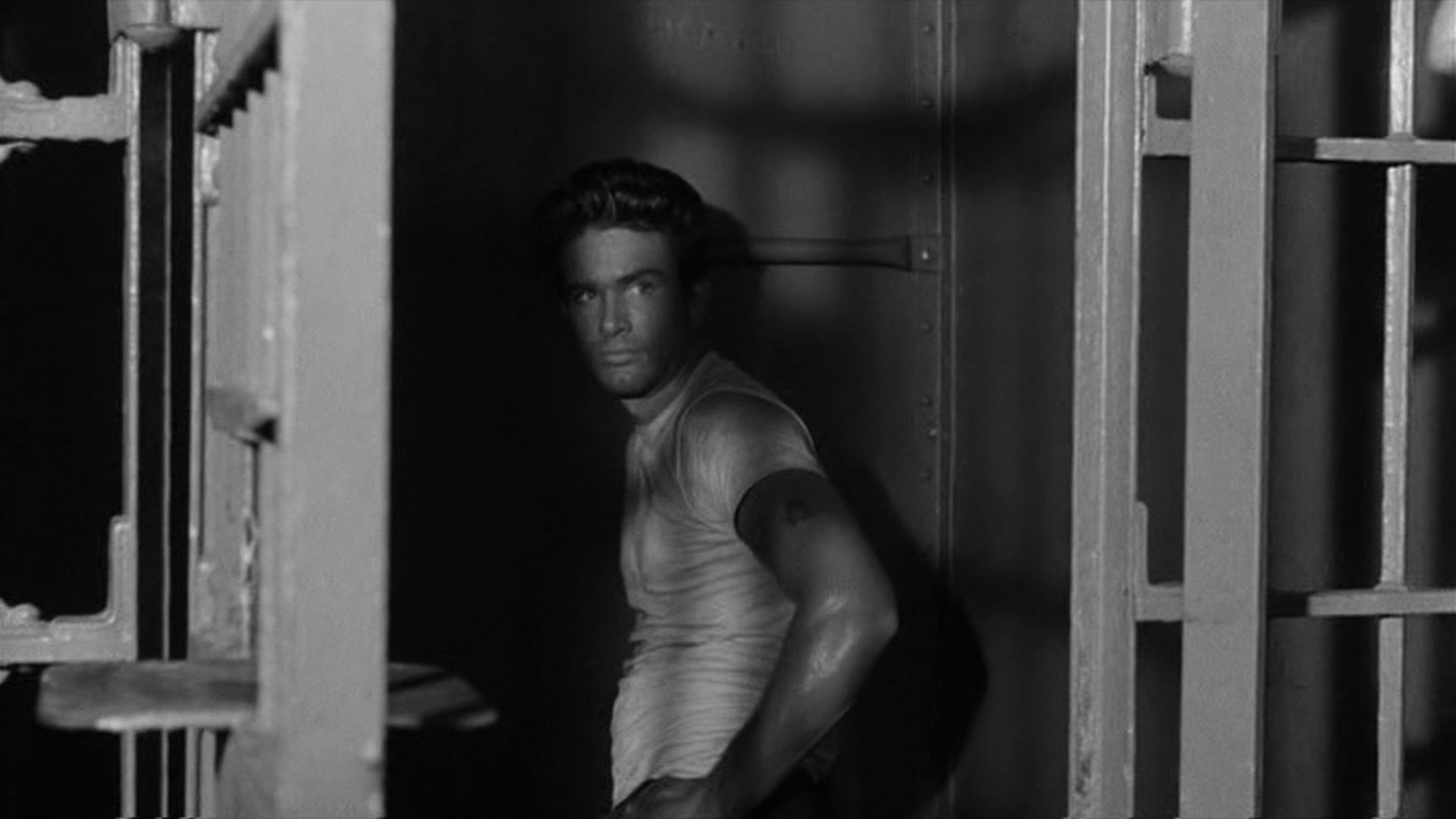 Warren Beatty blickt in trotziger Haltung und bei geöffneter Tür aus einer finsteren Gefängniszelle, deren Gitterstäbe Schatten auf sein weißes Unterhemd werfen.