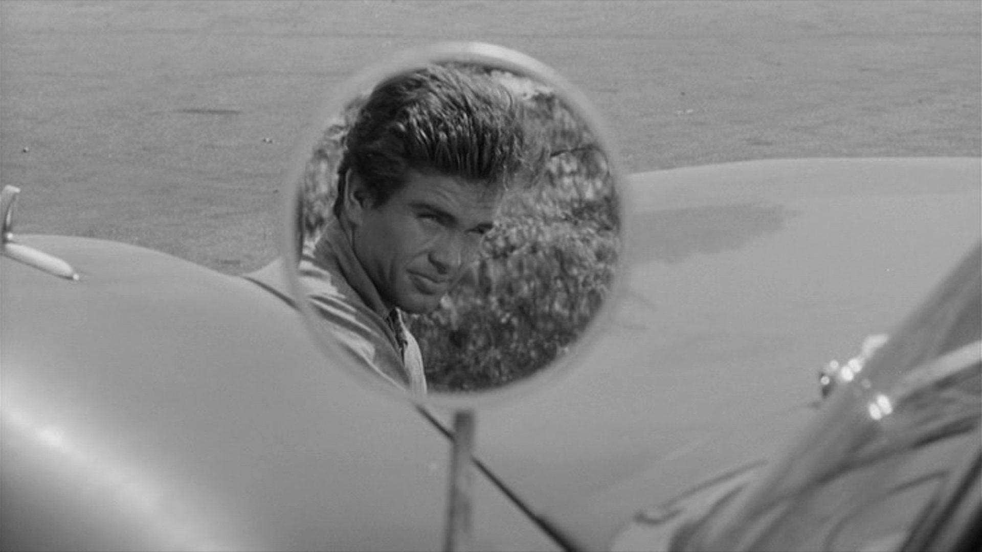 Schwarz-weiße Nahaufnahme eines runden Fahrzeugaußenspiegels, in dem sich Warren Beattys Kopf spiegelt.