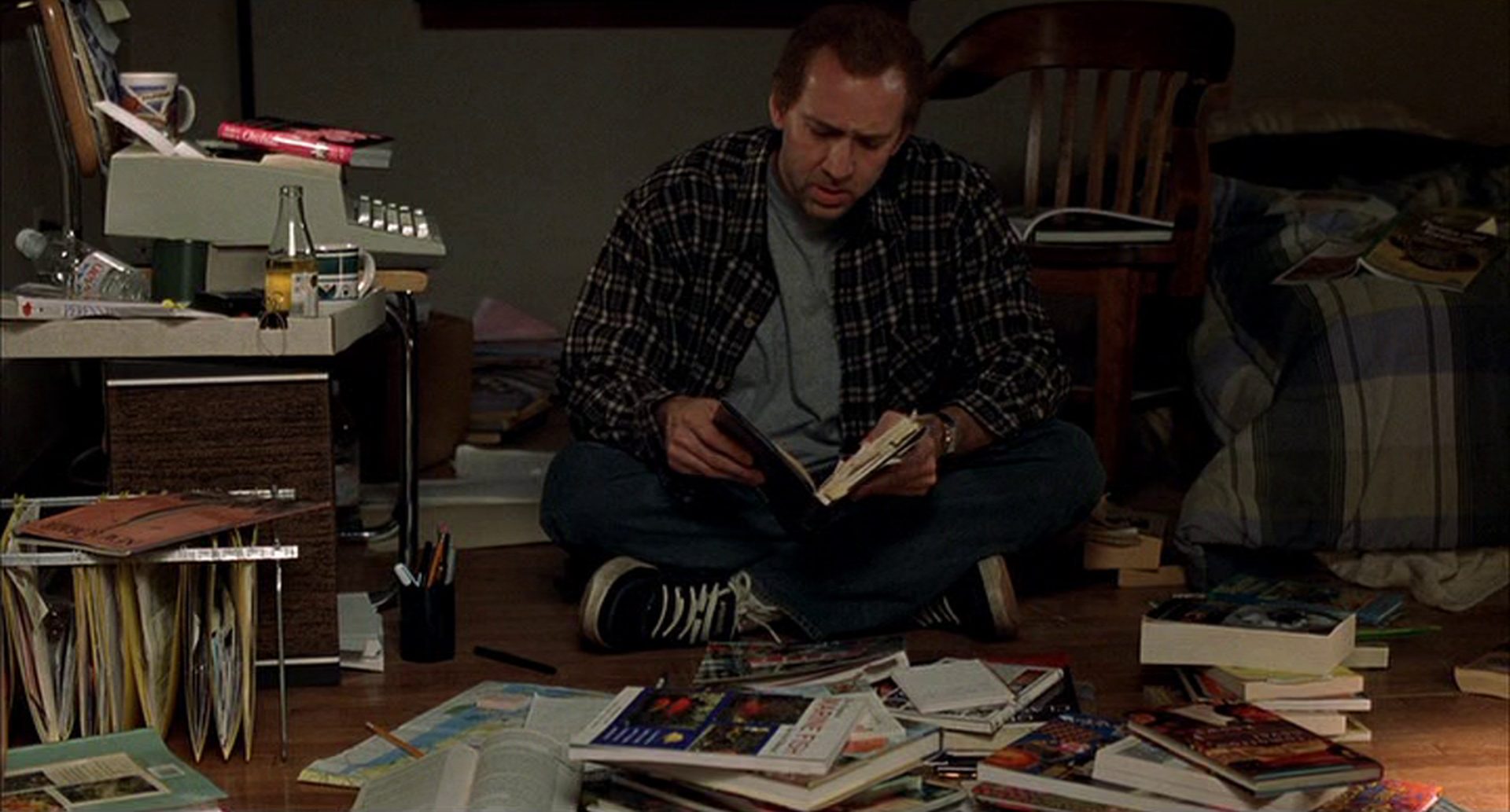 Aufnahme aus mittlerer Distanz von Nicolas Cage als Drehbuchautor Charlie Kaufman mit angestrengt-verzweifeltem Blick in ein Heft, im Schneidersitz auf dem Boden eines Zimmers in chaotischem Zustand, neben ihm auf einem Stuhl eine elektrische Schreibmaschine.