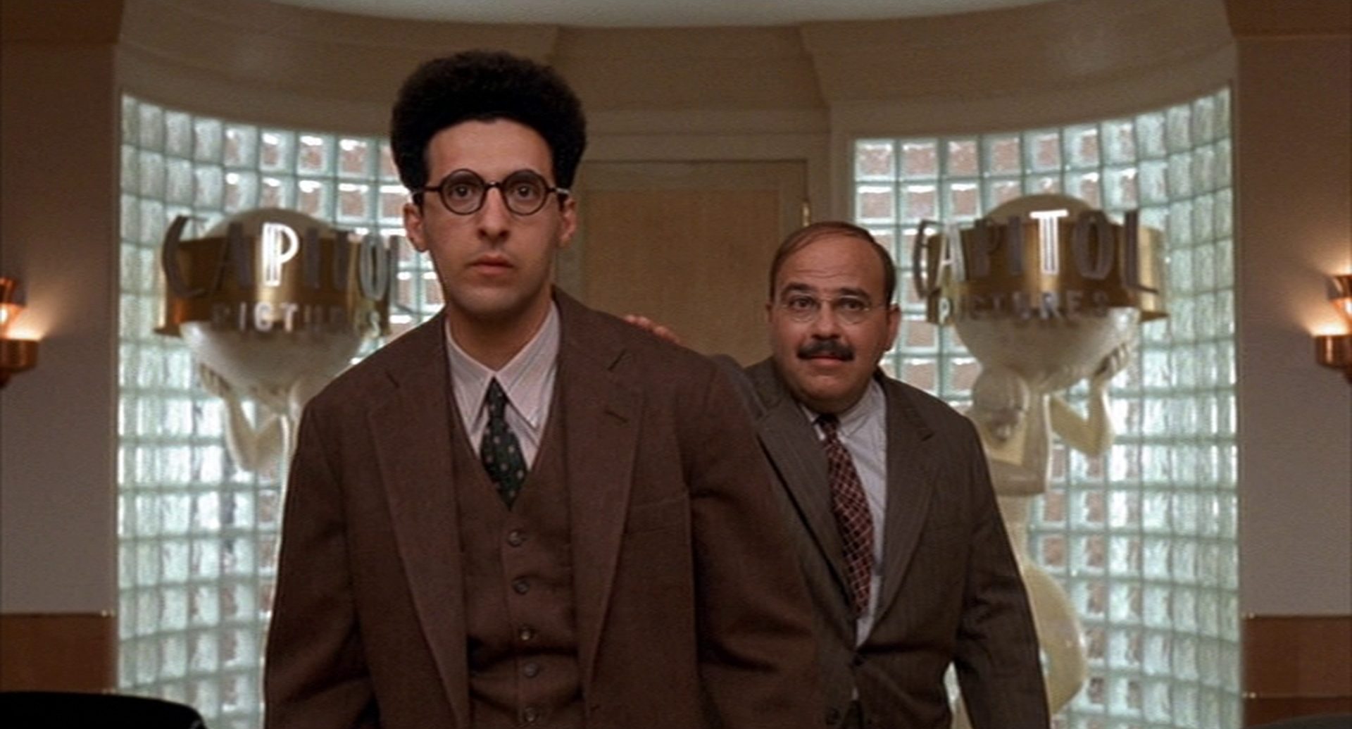 John Turturro als Barton Fink mit leicht perplexem Gesichtsausdruck im Büro des Studiobosses, im Hintergrund Jon Polito als Studiomitarbeiter.