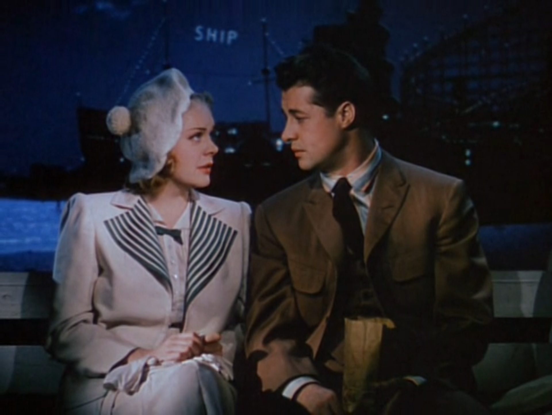 Technicolor-Aufnahme von Alice Faye als junge Schauspielerin Molly Adair und Don Ameche als junger Regisseur Michael Linnett Connors in abendlich-romantischer Atmosphäre auf einer Parkbank, im Hintergrund Küstenwasser und ein Schiff mit der Leuchtreklame Ship.