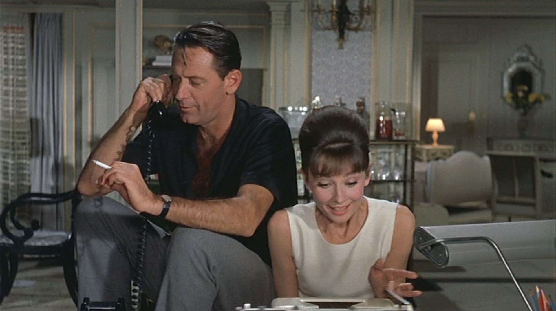 William Holden als legerer Drehbuchautor mit Zigarette in der Hand und Telefonhörer am Ohr in seinem Hotelzimmer; neben ihm sitzt Audrey Hepburn als Sekretärin an der Schreibmaschine; er sieht gelassen aus, sie enthusiastisch.