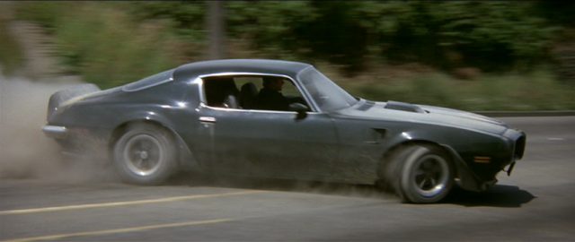 McQ schlittert in seinem Pontiac Firebird Trans Am mit rauchenden Reifen durch eine Kurve.