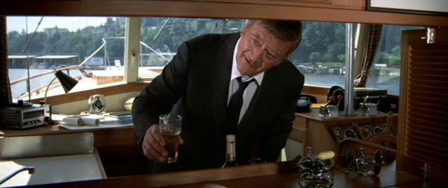 John Wayne als McQ auf seiner Jacht mit einem Drink in der Hand.