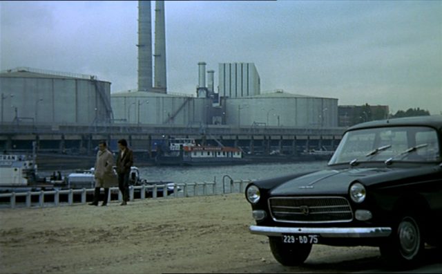 Blick auf eine triste Industriekulisse; im Hintergrund gehen zwei Männer durch die Szenerie, im Vordergrund parkt ein dunkles Auto.