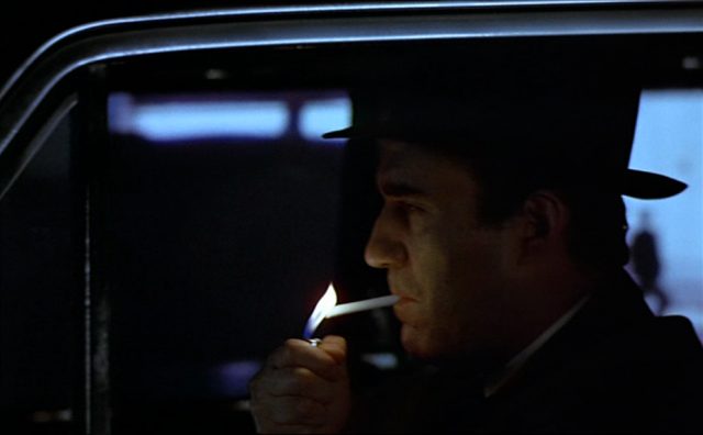 Seitliche, dunkle Nahaufnahme von Max am Steuer seines Fahrzeugs, wie er sich gerade eine Zigarette anzündet.