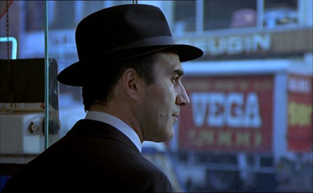 Close-up von Michel Piccoli als Kriminalkommissar Max mit Hut beim Blick auf eine großstädtische Straße.