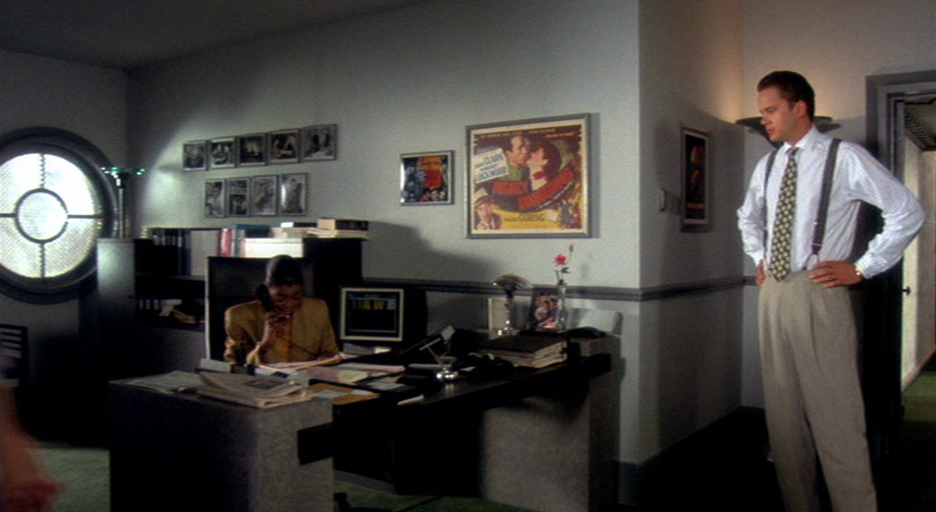 Griffin Mill (Tim Robbins) steht ohne Sakko mit Krawatte und Hosenträgern vor dem Schreibtisch seiner Sekretärin (Angela Hall), die gerade telefoniert.