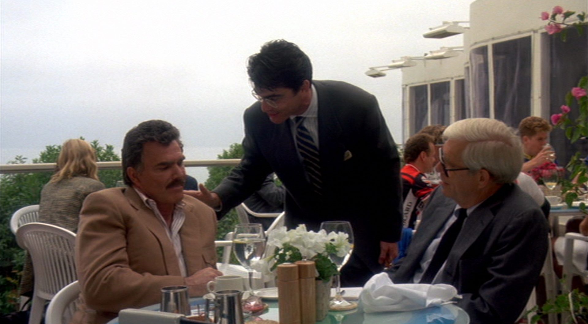 Studioproduzent Larry Levy (Peter Gallagher) begrüßt auf der Terrasse eines Edelrestaurant Burt Reynolds, der sich an einem Tisch unterhält.