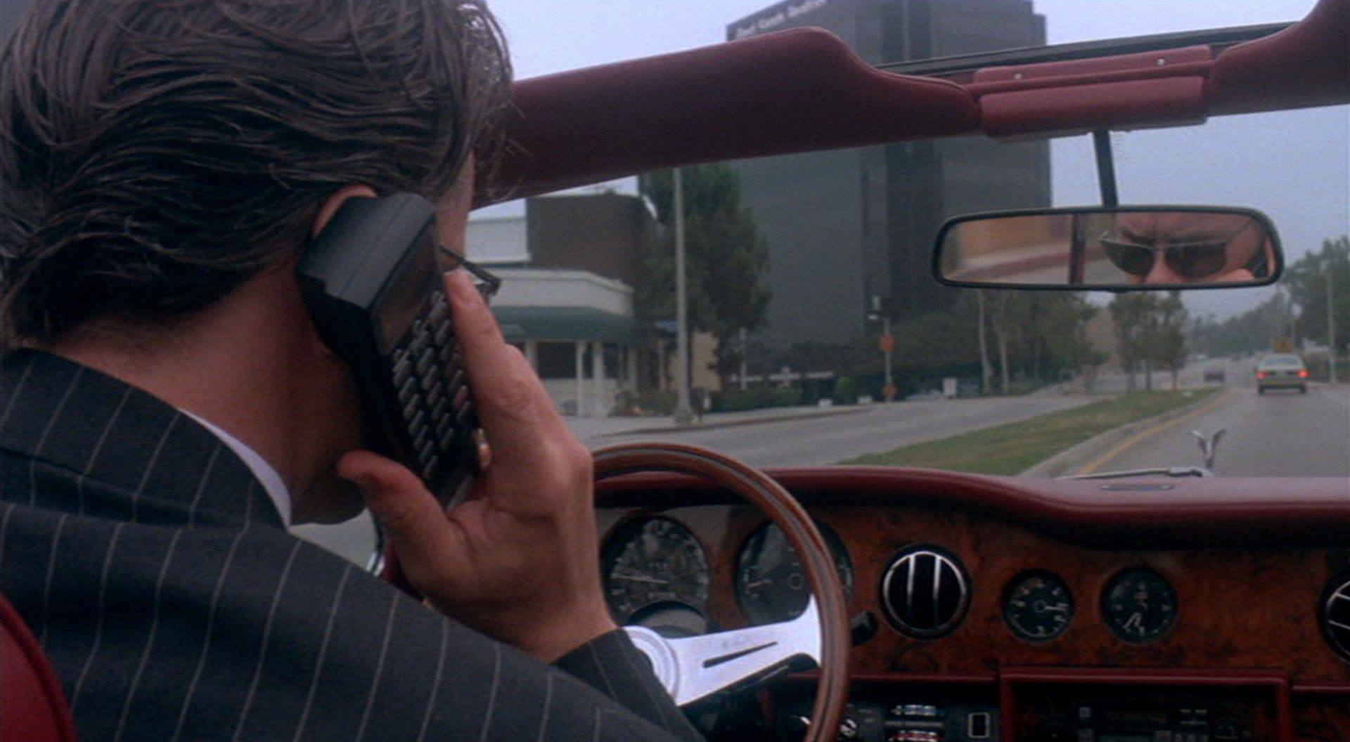Blick von hinten auf Griffin Mill (Tim Robbins) am Steuer seines Luxuscabriolets auf der Fahrt durch Los Angeles, am Ohr den Hörer seines Autotelefons; im Rückspiegel sieht man eine hinter seiner Sonnenbrille verborgene Gesichtspartie.