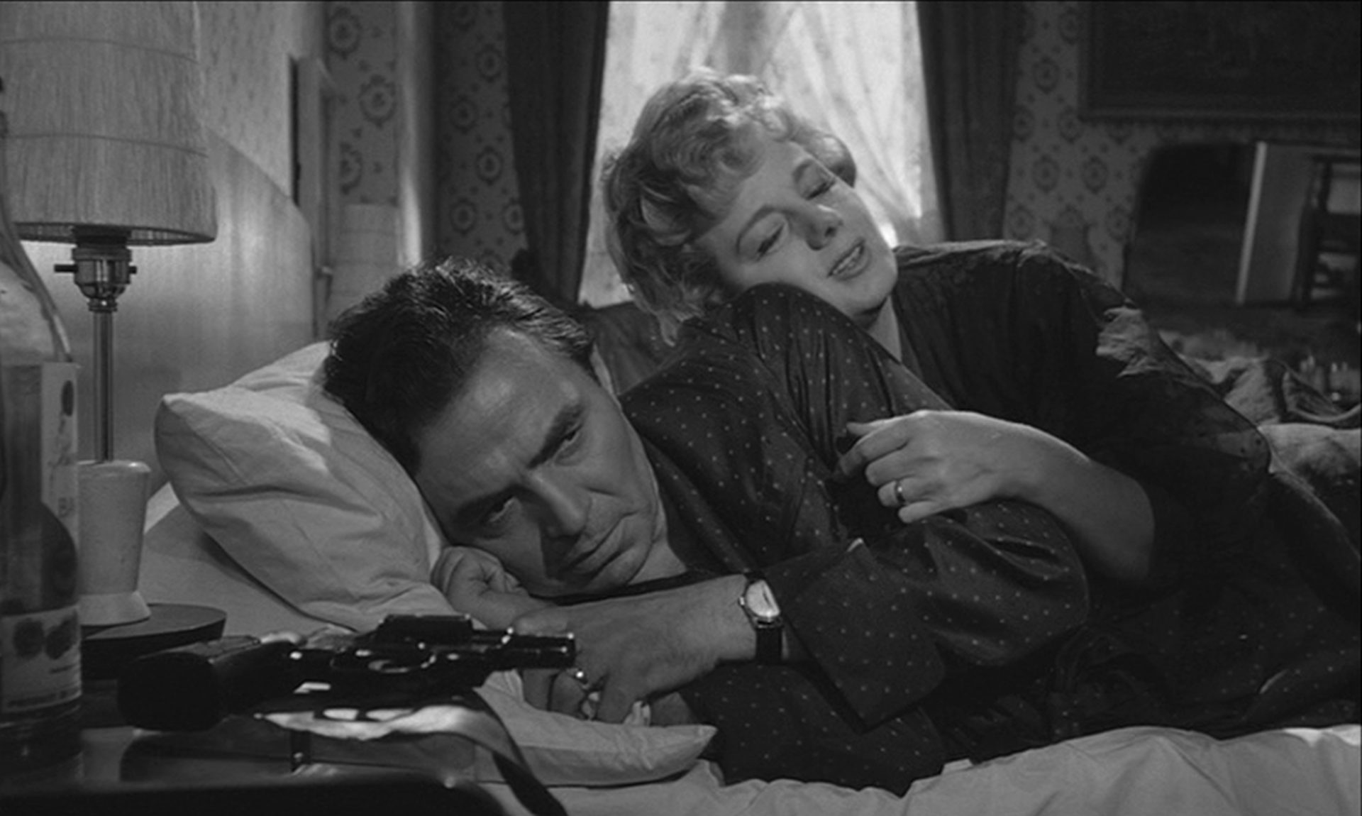 James Mason als Humbert Humbert und Shelley Winters als Charlotte Haze; beide liegen im Bett. Während ihr Gesichtsausdruck romantisches Wohlbefinden verheißt, blickt er genervt drein. Auf dem Nachttisch liegt ein Revolver.