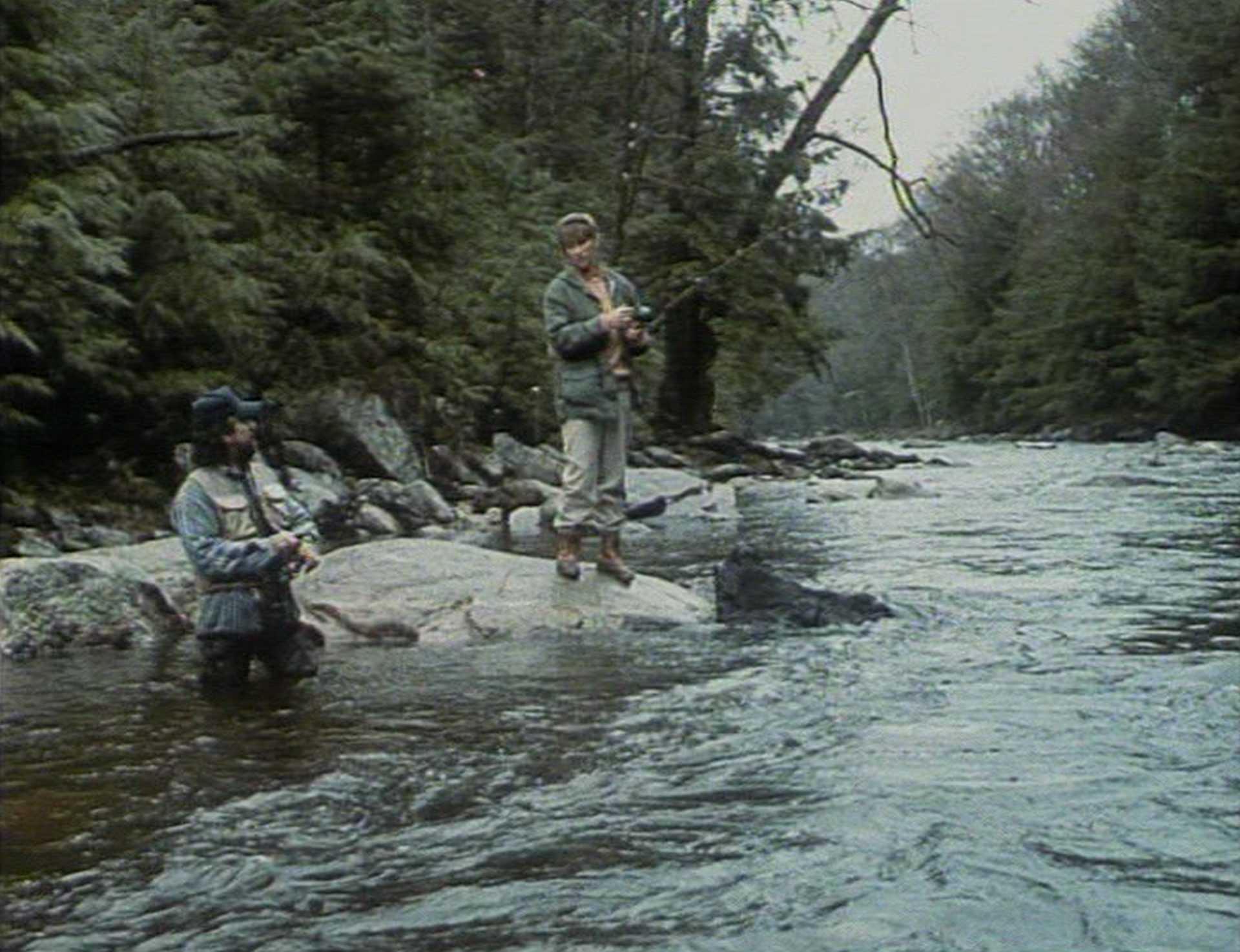 Zwei Personen beim Angeln in der Wildnis, eine steht im Fluss, die andere auf einem Fels.
