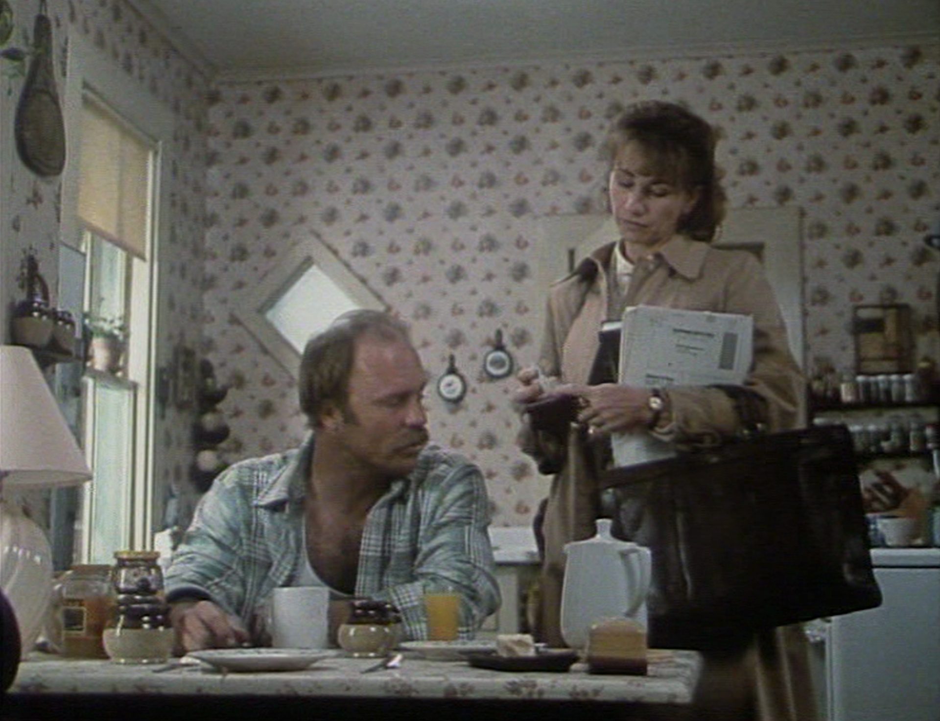 Ed Harris mit Schnäuzer und Kathy Bates in einer proletarisch anmutenden Küche; er sitzt am Tisch, während sie im Mantel und mit Aktentasche neben ihm steht.
