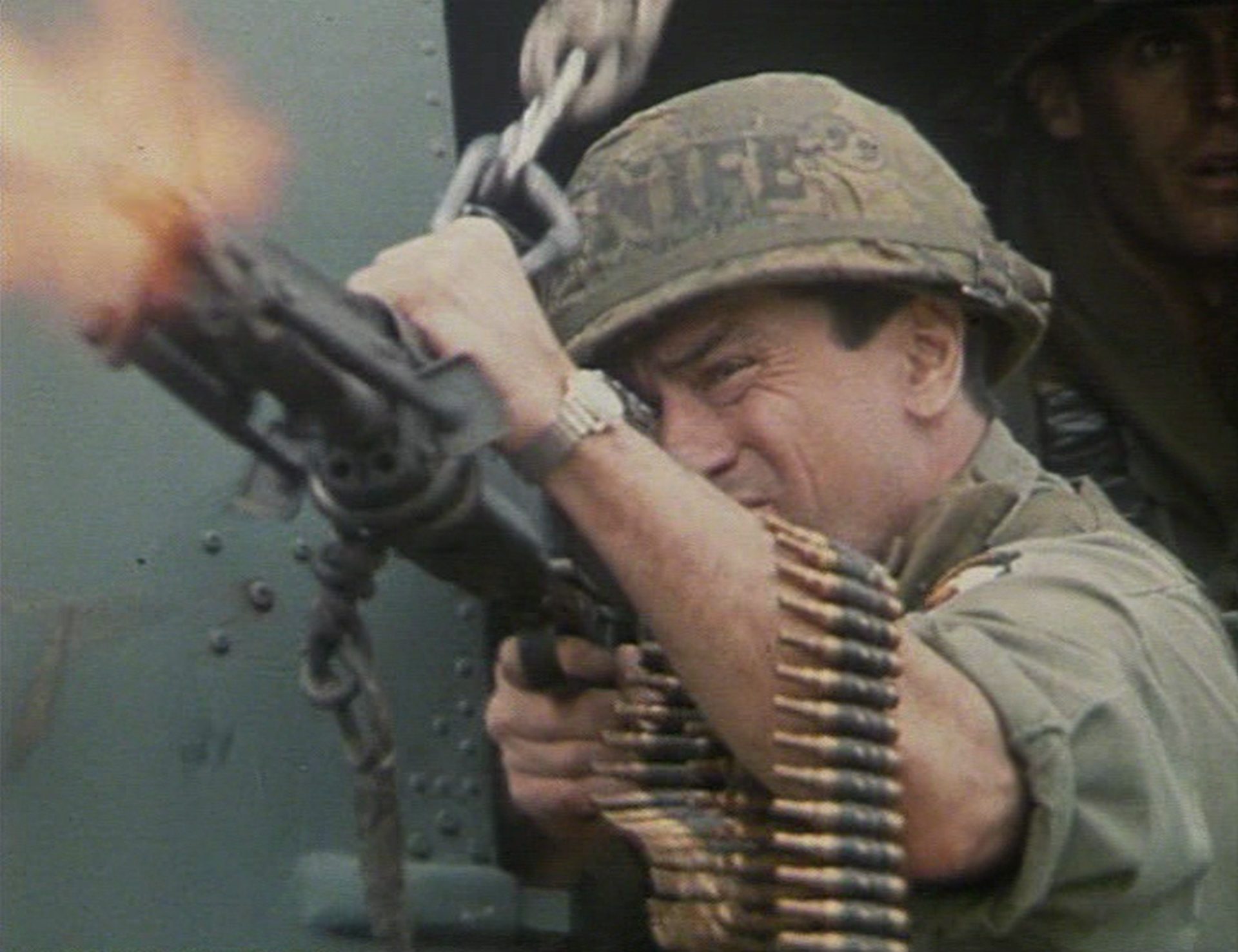 Nahaufnahme von Robert De Niro in Soldatenmontur beim Abfeuern eines Maschinengewehrs, einen Patronengurt um den Unterarm gewickelt.
