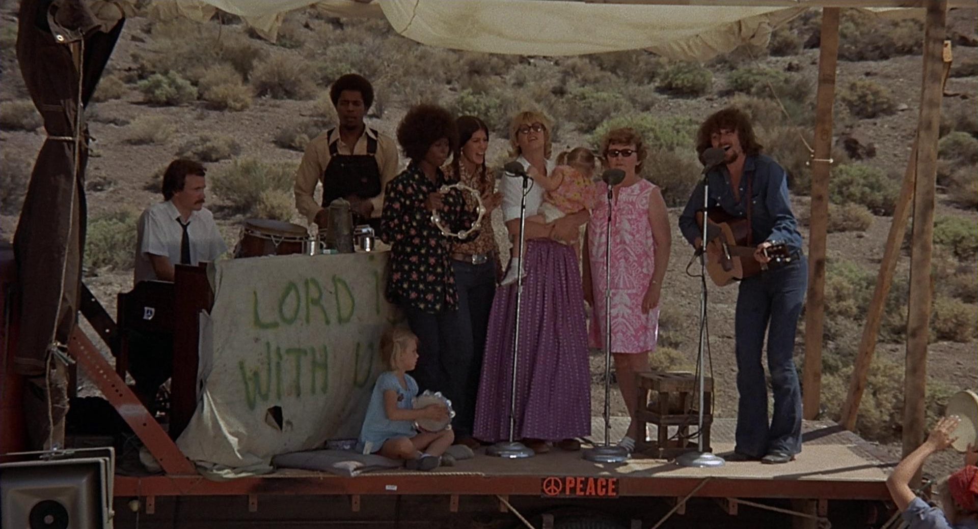 Szene mit gegenkulturellem Flair: Auf einer provisorischen Bühne mitten in der Wüste musizieren Menschen unterschiedlicher Hautfarbe und Altersgruppen.