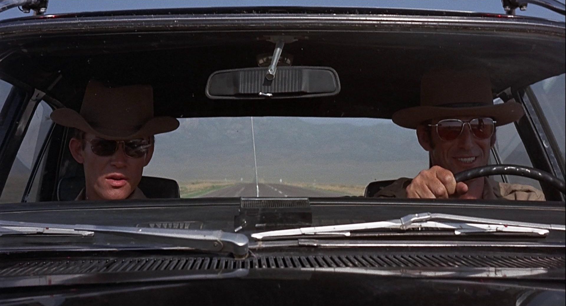 Frontale Nahaufnahme von zwei sonnenbebrillten Polizisten in ihrem Streifenwagen, im Hintergrund zeichnen sich am Horizont Berge in einer unbewohnten Gegend ab; der Fahrer lächelt.
