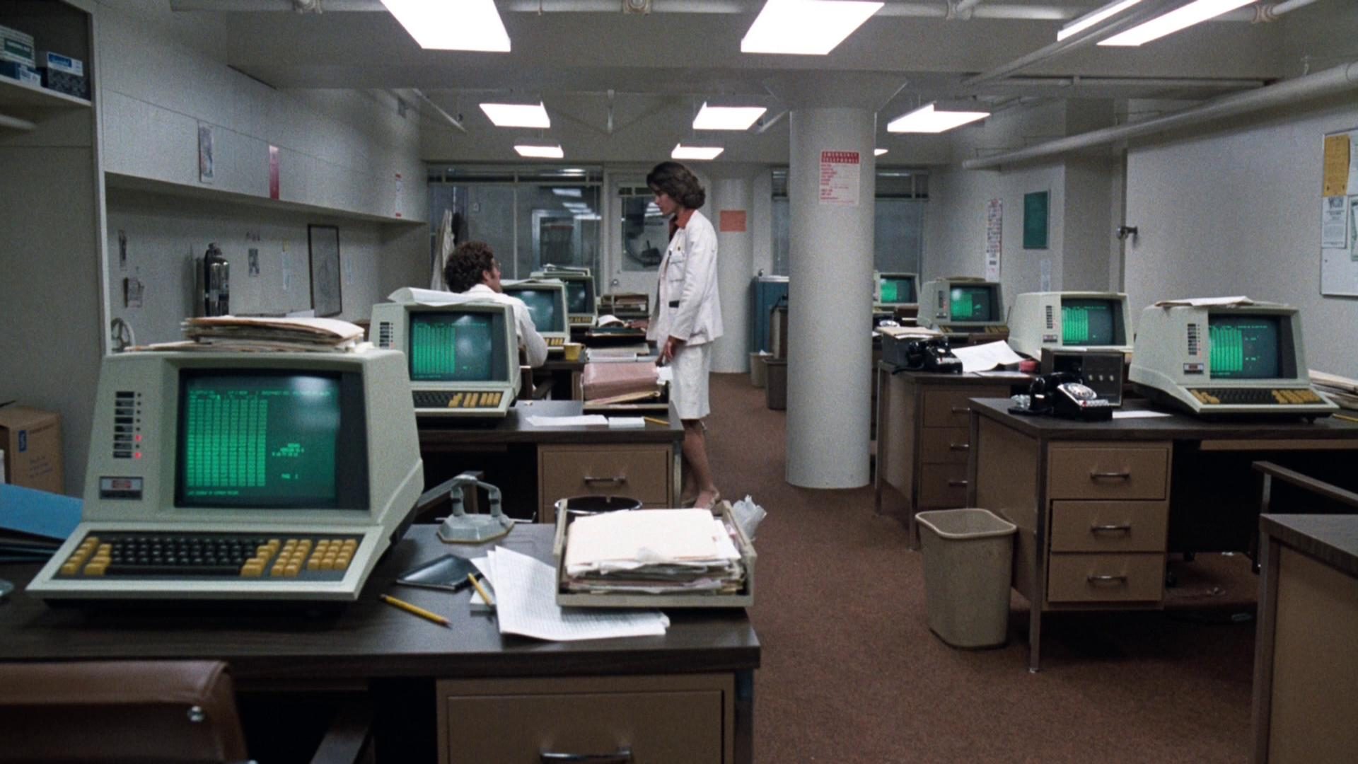 Blick in einen Krankenhausraum voller Arbeitsplätze mit Computern auf dem technischen Stand der 1970er Jahre, eine Ärztin unterhält sich mit einem Kollegen.