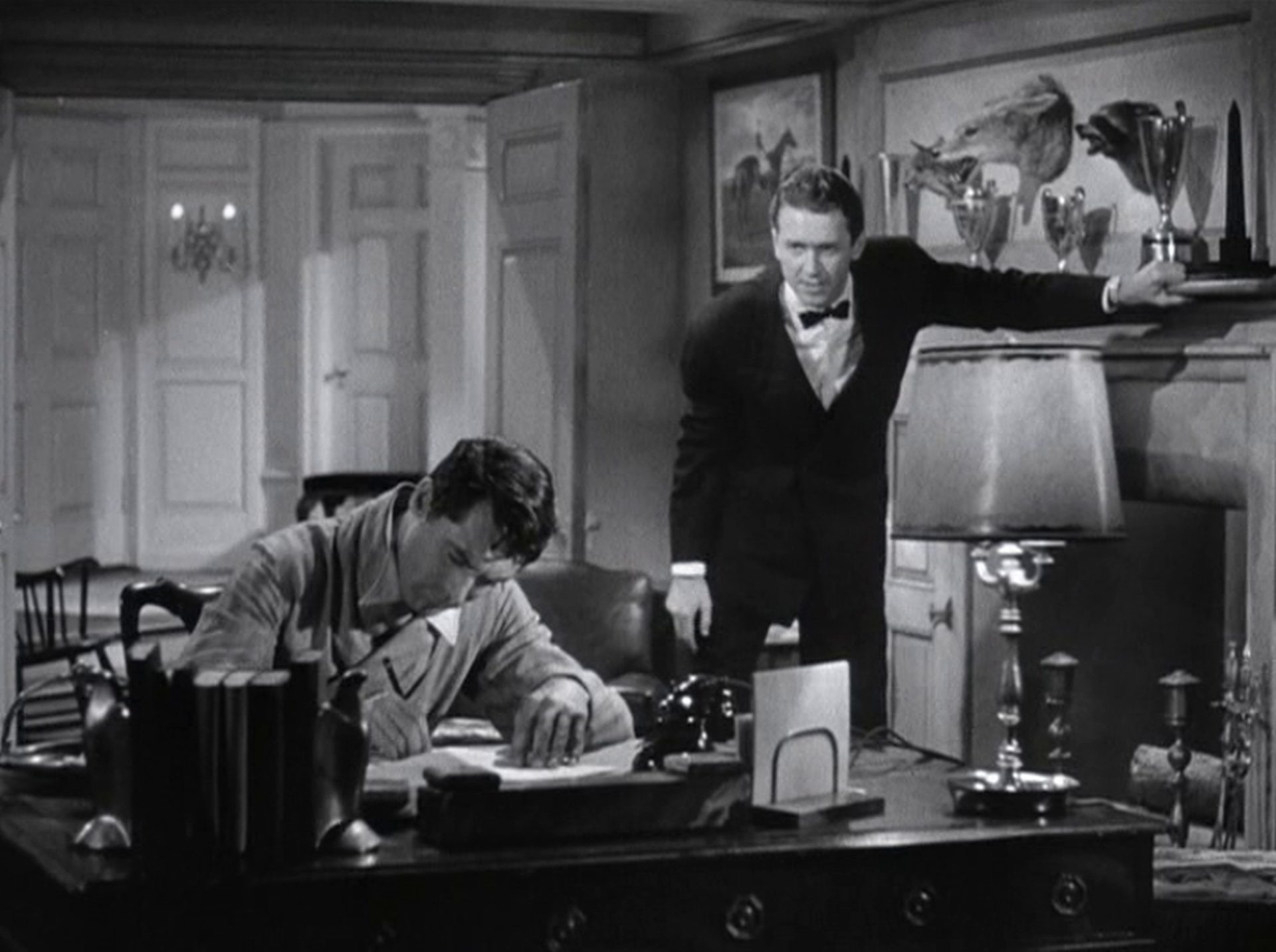 Cary Grant notiert etwas am Schreibtisch, neben ihm im Hintergrund James Stewart in erschöpfter Körperhaltung.