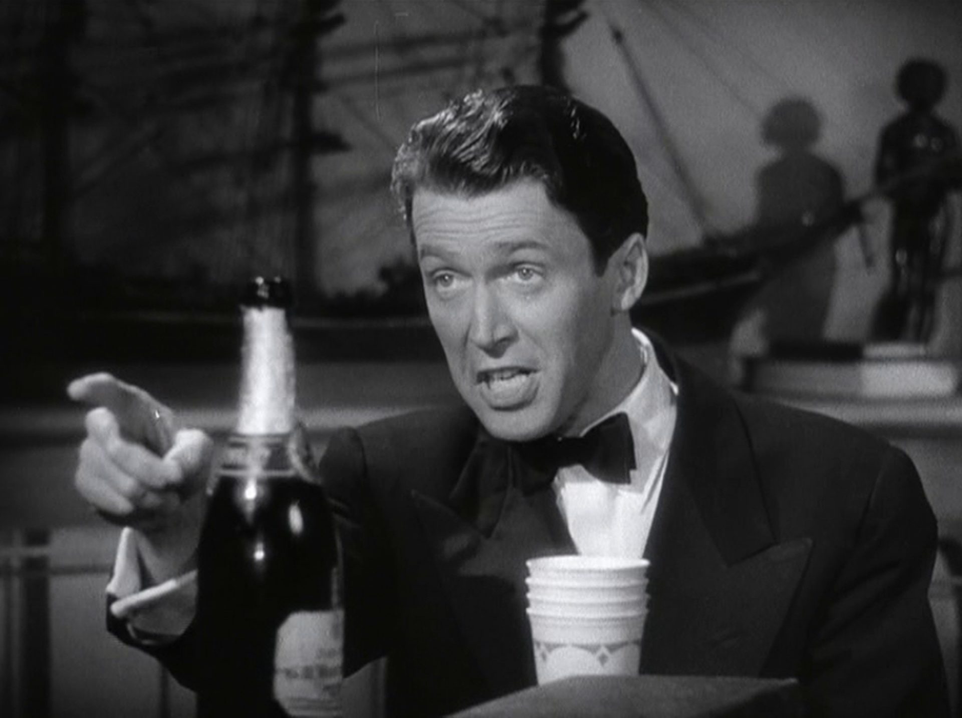 Nahaufnahme von James Stewart als Connor in Abendkleidung, vor ihm eine geöffnete Champagnerflasche in angeregter Unterhaltungspose.