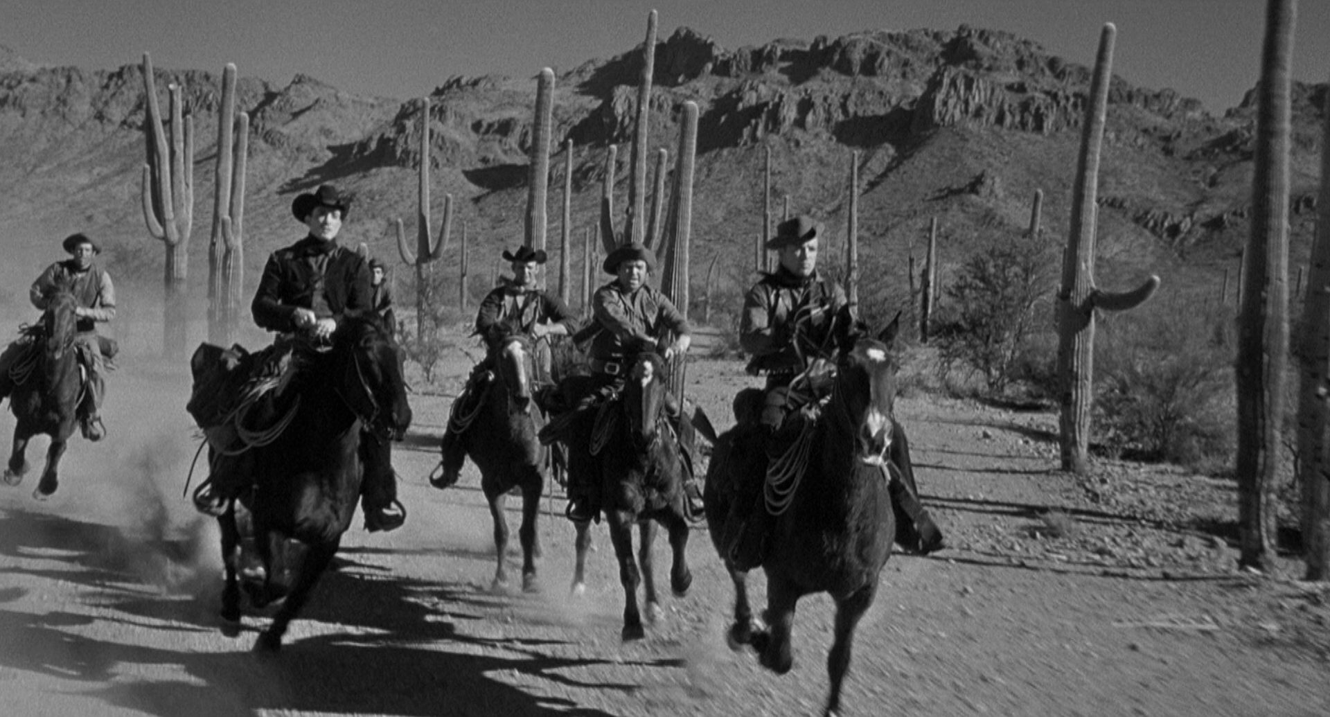 Fünf Banditen reiten durch eine Kakteenlandschaft, im Hintergrund staubige Gebirge.