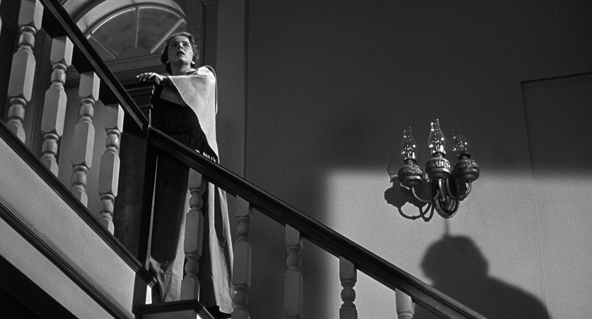 Leora Dana als Alice Evans auf der Hoteltreppe; sie starrt mit schockiertem Blick in Richtung Gebäudedecke, ein Schatten rechts unten im Bild deutet einen aufgeknüpften Mann an.
