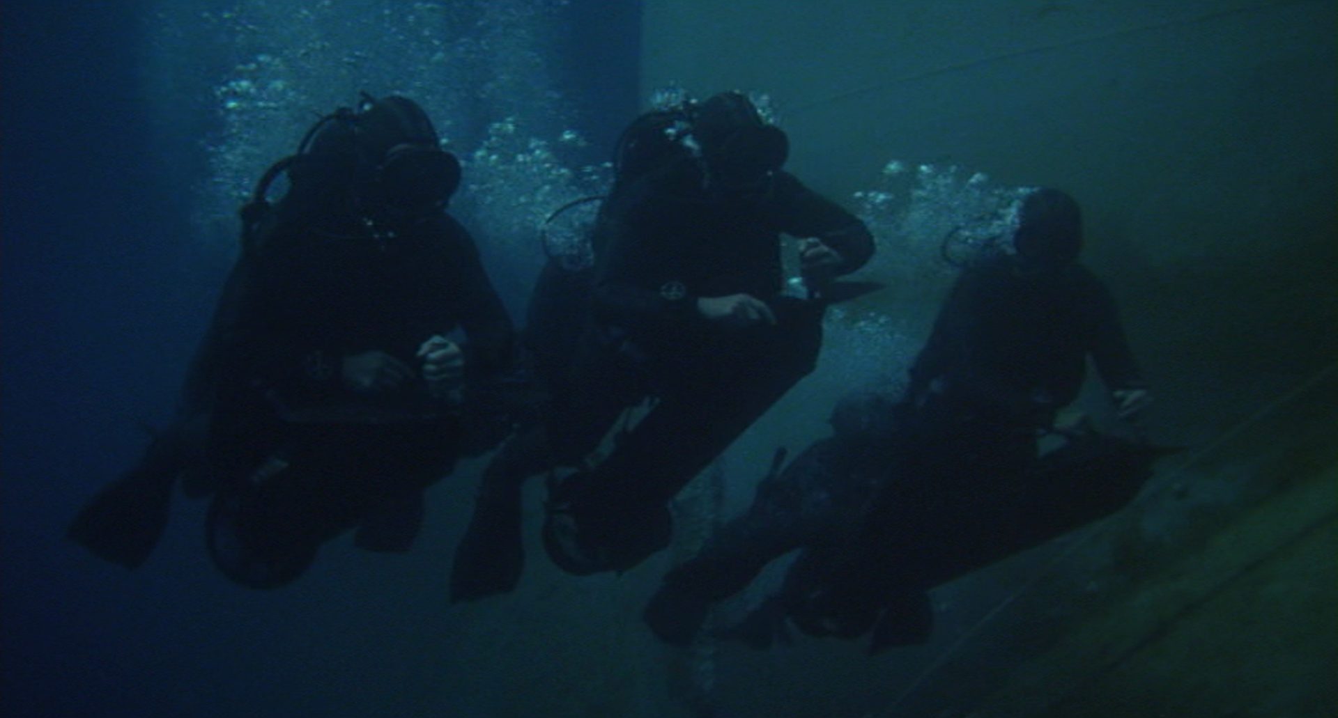 Drei Taucher in dunklen Anzügen unter Wasser.