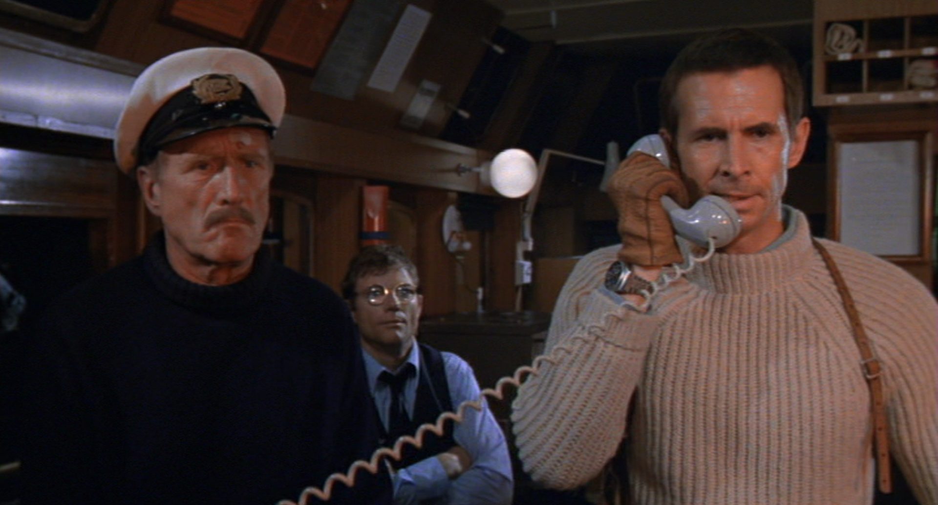 Anthony Perkins als überlegen dreinblickender Terrorist Kramer im Rollkragenpullover bei einem Telefonat; neben ihm ein Seemann, im Hintergrund ein Komplize.