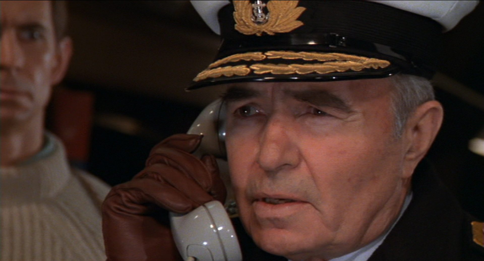 Nahaufnahme von James Mason als Admiral Brinsden mit ernster Miene mit einem Telefonhörer am Ohr; im Hintergrund ist Kramer mit finsterem Blick erkennbar.