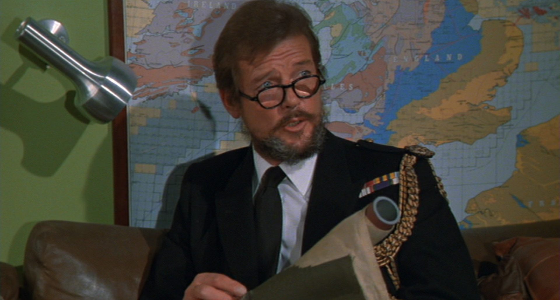 Roger Moore, mit Vollbart und Hornbrille, als ffolkes in exzentrischer Pose vor einer Karte.