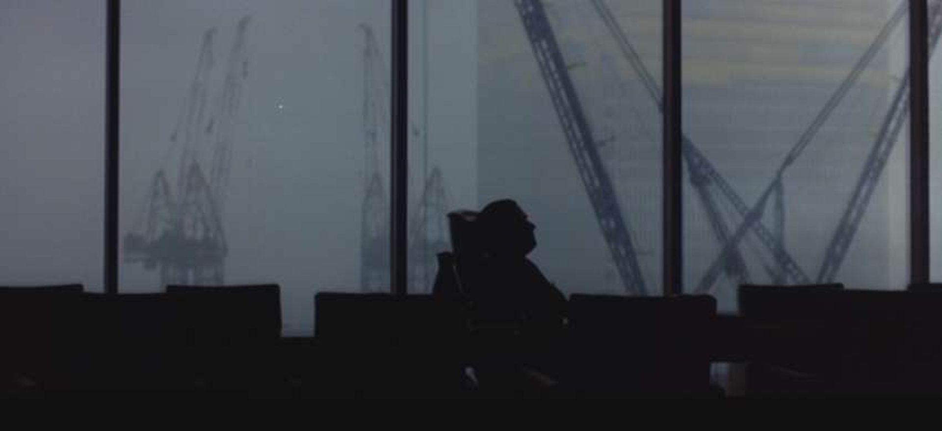 Schwarze Silhouette eines Mannes, der in seinem Bürostuhl vor einem Panoramafenster lehnt; draußen sind mehrere Kräne und ein Hochhaus erkennbar.