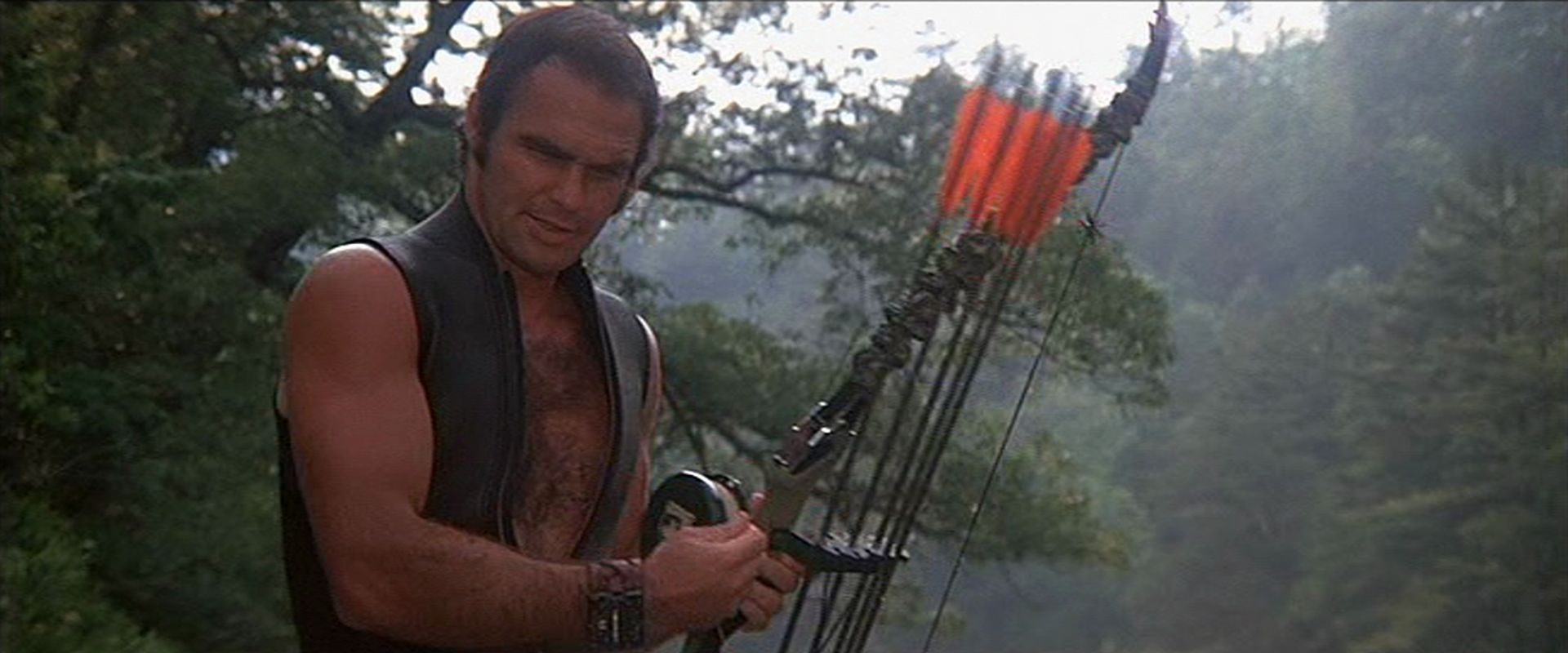 Burt Reynolds in konzentrierter Pose als Abenteurer in ärmelloser Neoprenweste und mit Bogen in der Wildnis.