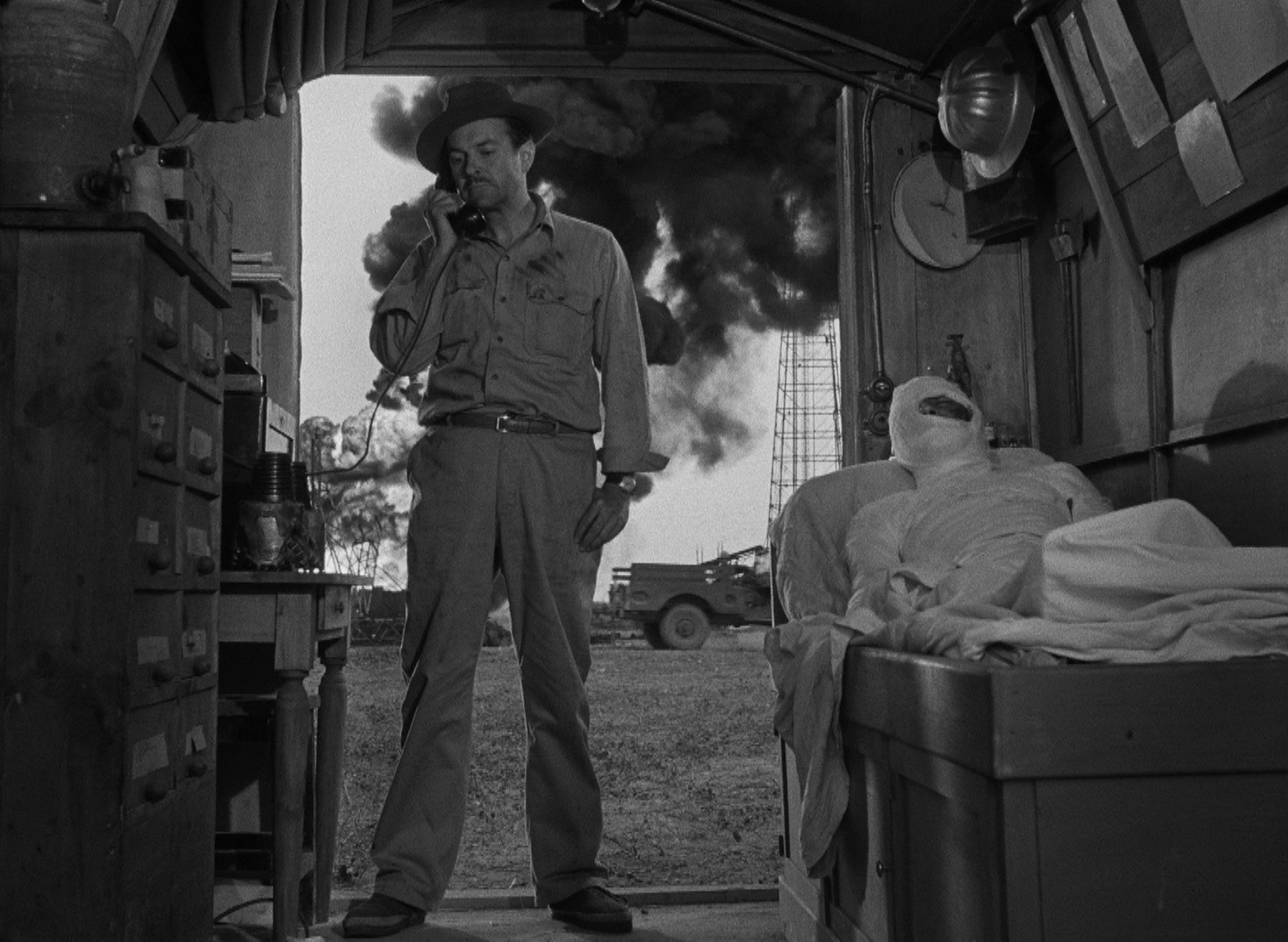 Ölarbeiter am Telefon in einer kleinen Hütte, neben ihm ein Mann im Ganzkörperverband, im Hintergrund brennt ein Bohrloch mit riesigen Flammen und einer dunklen Qualmwolke.