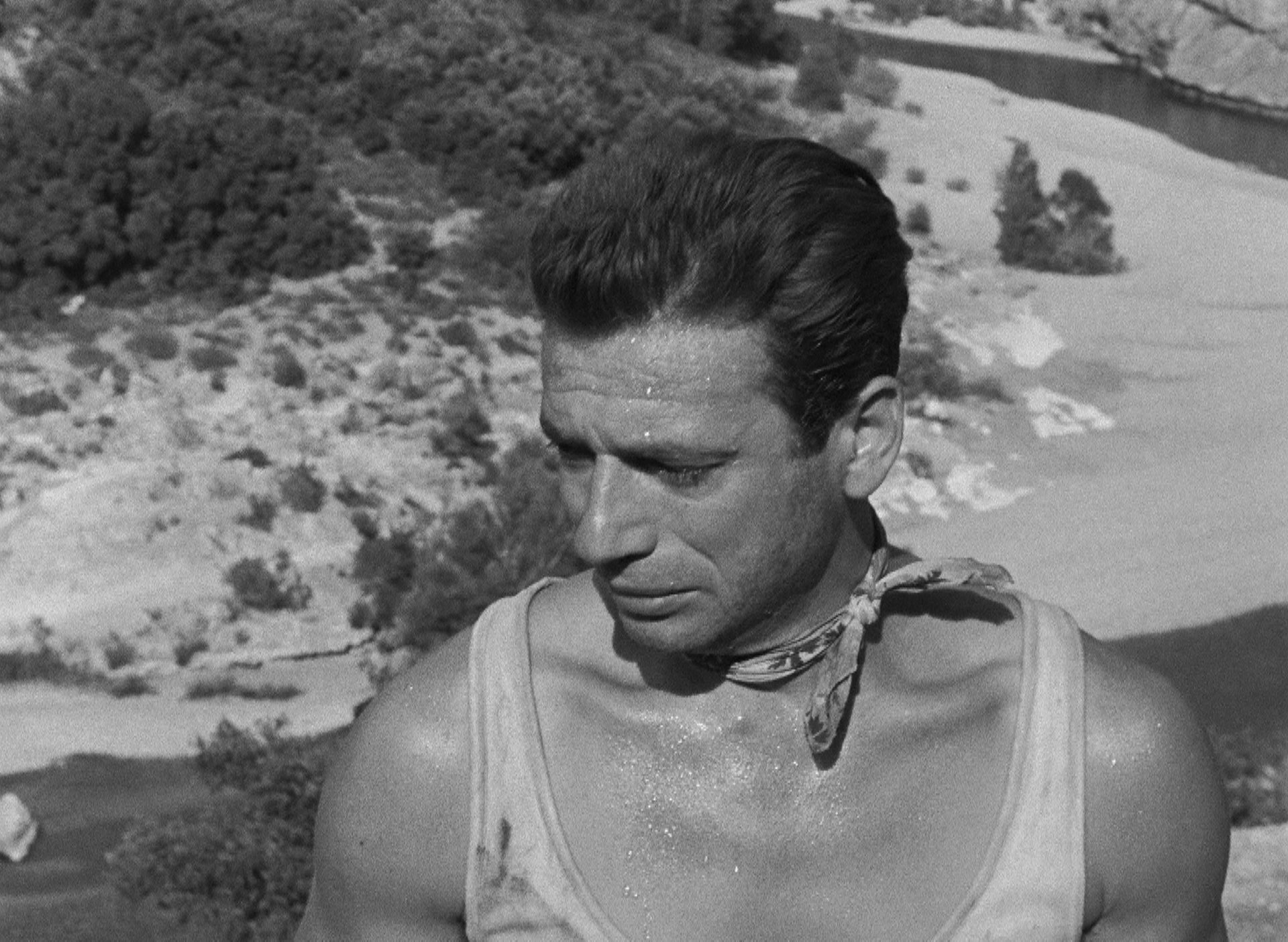 Nahaufnahme von Yves Montand mit Schweißperlen auf Stirn und Brust vor dem Hintergrund einer Hügelkette.