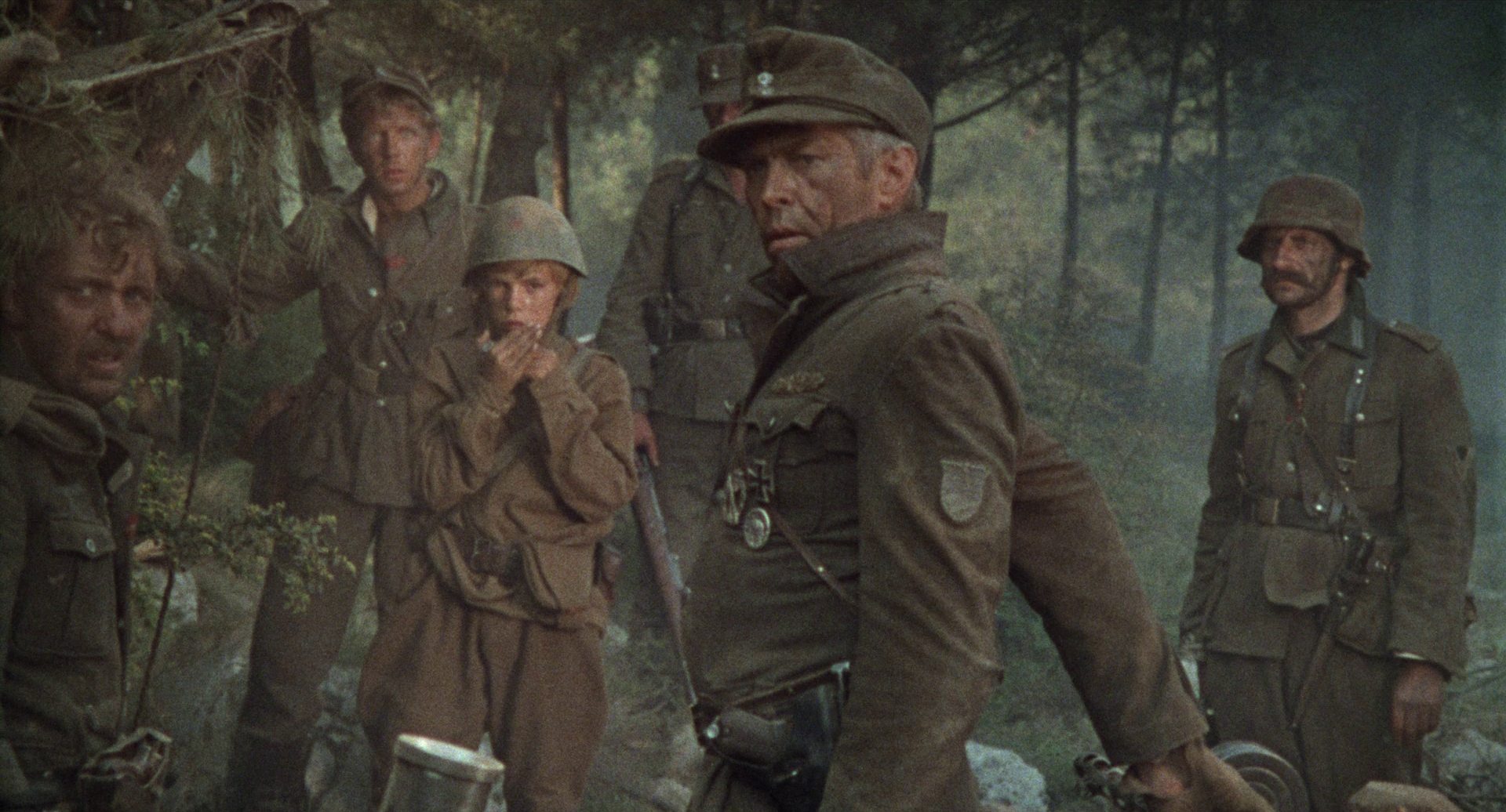James Coburn als Steiner im Beisein seiner Leute mit einem kriegsgefangenen Kindersoldaten, der Mundharmonika spielt, am Waldrand.